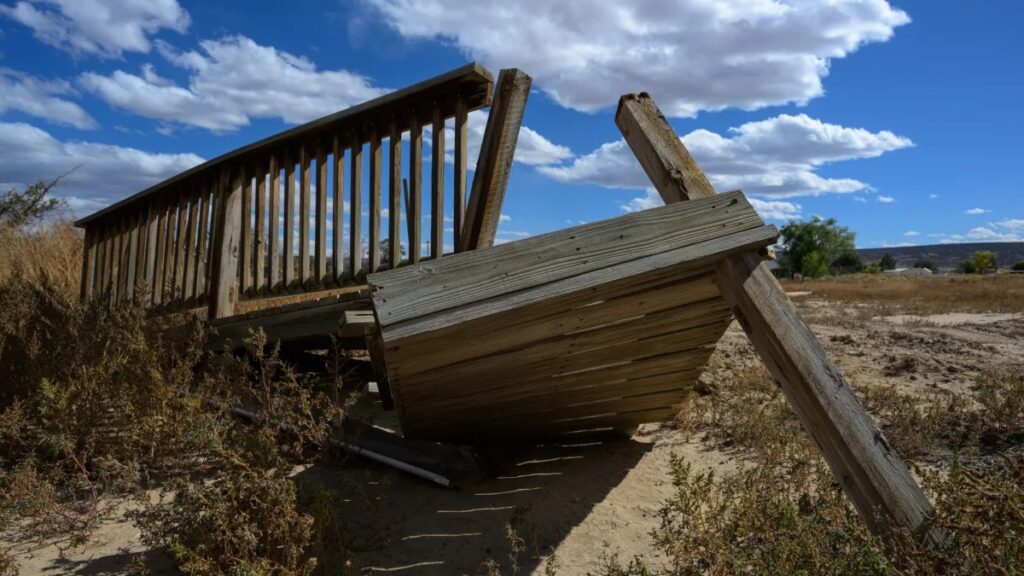 A broken wooden bridge lies collapsed on a dirt path, under a blue sky with scattered clouds. Surrounding the bridge are dry grass and sparse vegetation.