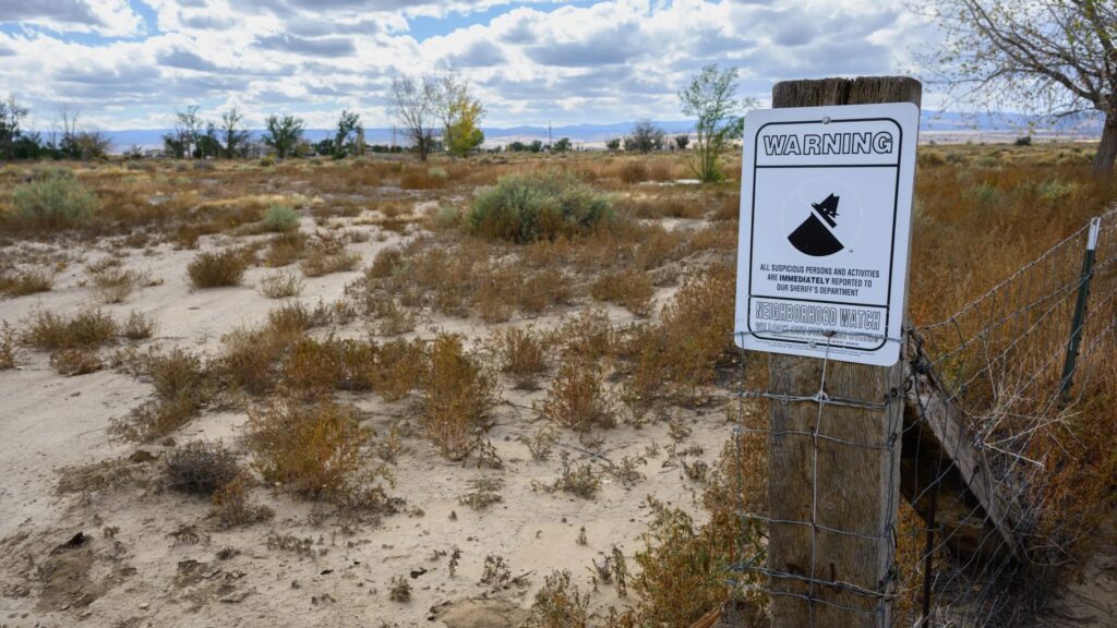 A sign on a fence warns of surveillance in a dry, sparse landscape with scattered shrubs and a partly cloudy sky.