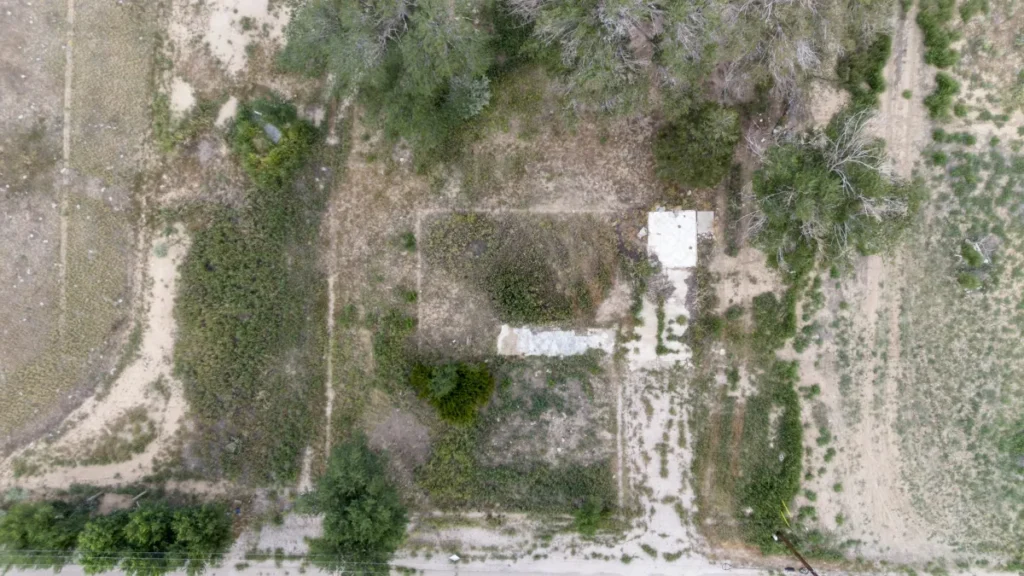 Aerial view of a grassy area with scattered trees and vegetation. A concrete slab is visible on the right side, with paths and dirt patches throughout.