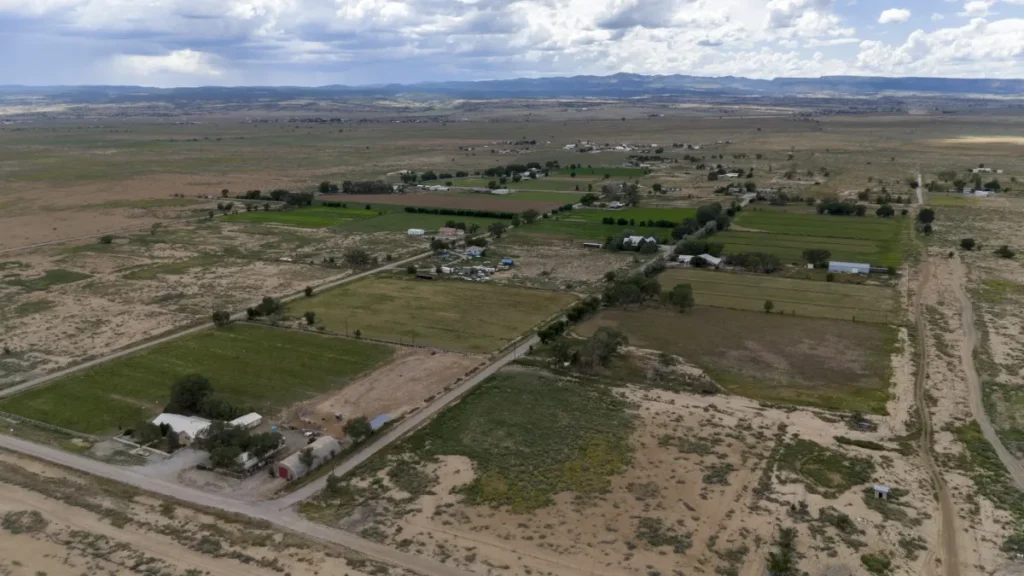 Aerial view of a rural landscape with scattered buildings, patches of green fields, and dry sandy areas under a cloudy sky.