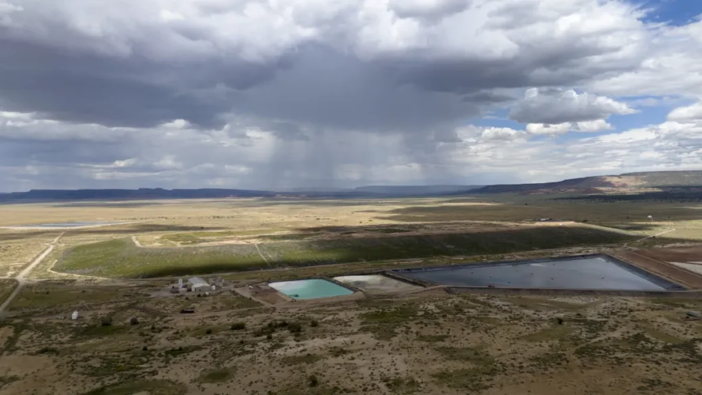 Aerial view of a desert landscape with two rectangular water bodies, cloudy sky, and distant rain showers. Sparse vegetation and a few structures are visible.