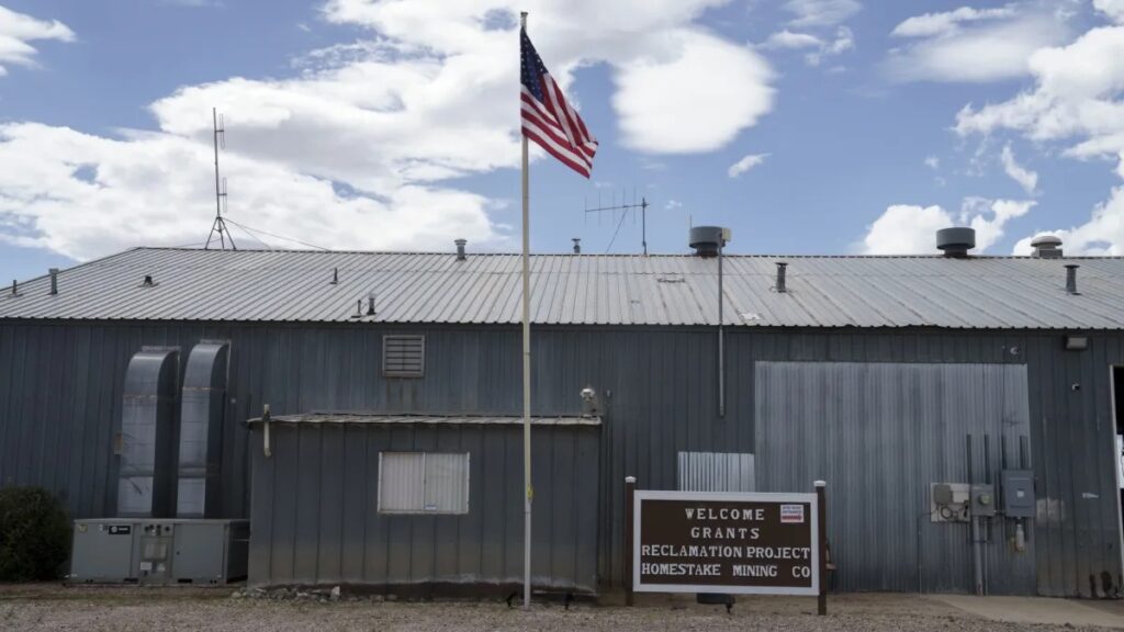 A building with a metal roof and an American flag in front. A sign reads, "Welcome Grants Reclamation Project Homestake Mining Co." The sky is partly cloudy.