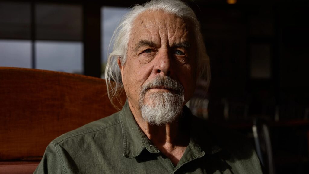 Older man with long white hair and a beard, wearing a dark green shirt, sits in a dimly lit room, gazing forward with a serious expression. Shadows fall across his face.