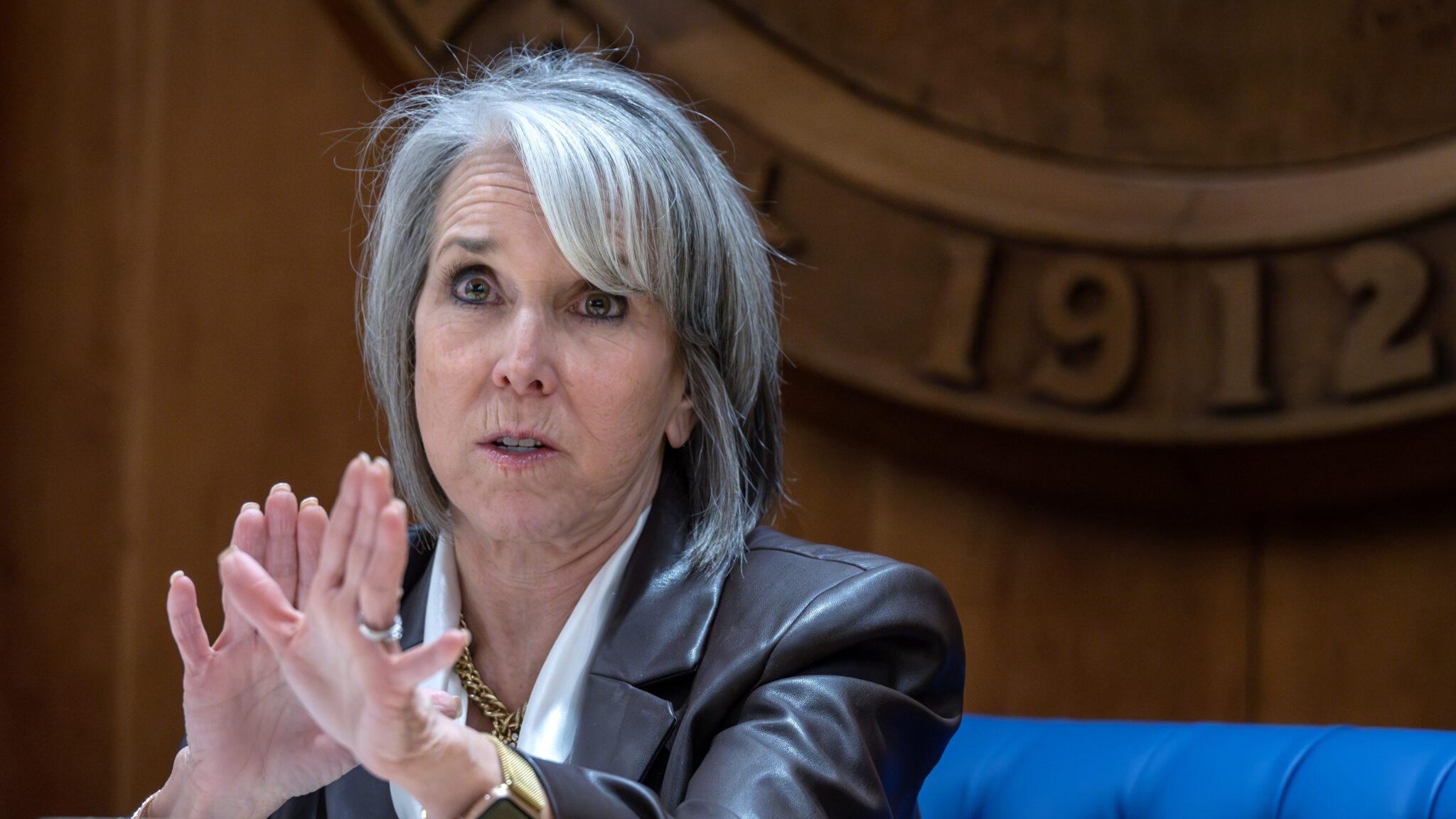 A person with gray hair wearing a dark blazer gestures with hands while speaking indoors, in front of a wooden emblem.