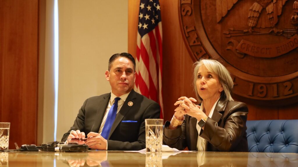 Two individuals sit at a conference table with an American flag and a large emblem in the background. The woman is speaking, and the man is looking forward. Glasses of water are on the table.