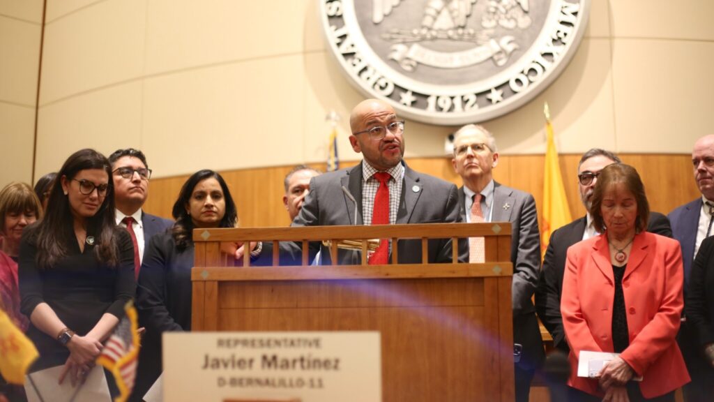 A person stands at a podium in a formal room, surrounded by others. A nameplate with "Javier Martinez" is visible. Several flags are in the background.