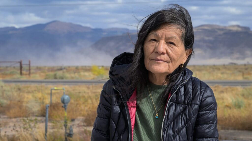 A woman with long black hair and a black jacket stands outdoors in a windy, mountainous landscape.