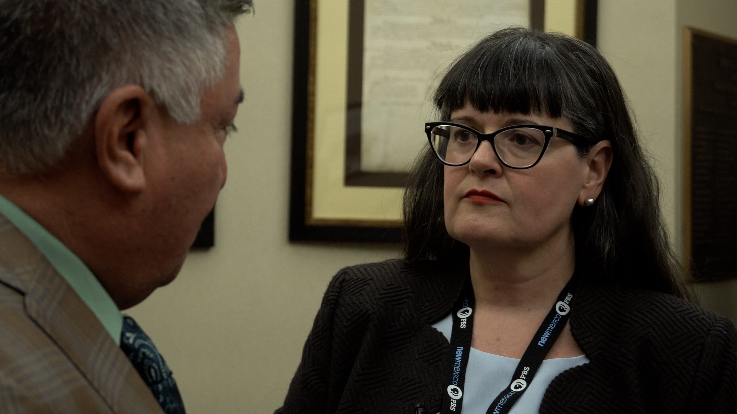 A woman with glasses and a lanyard listens intently to a man in a suit. Framed documents are visible on the wall behind them.