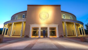 Modern government building with a round structure, large entrance, and seal above the doorway. Lit by exterior lighting against a clear evening sky.