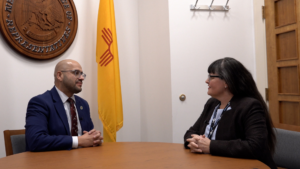 Two people seated at a table in an office with a New Mexico flag and a seal on the wall.