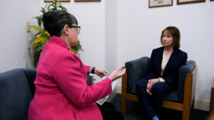 Two women are sitting in an office, engaged in conversation. The woman on the left is gesturing with her hands, while the woman on the right listens attentively.