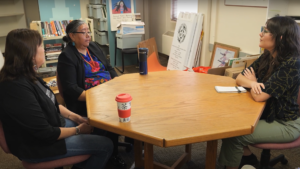 Three people seated around a wooden table in an office setting, engaged in conversation. Various office supplies and posters are visible in the background.