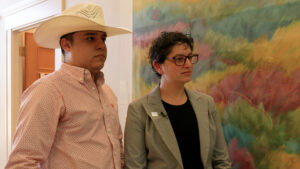 Two people stand side by side indoors. One wears a cowboy hat and patterned shirt, the other wears glasses and a suit. They are in front of a colorful abstract painting.