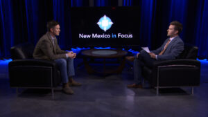 Two people are seated facing each other in a television studio. A screen behind them displays the text "New Mexico in Focus.