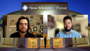 Two people are on a split-screen video call during a "New Mexico in Focus" segment. Both wear glasses and face the camera against a backdrop of framed pictures and a blurred building exterior.