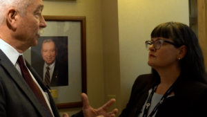 Two people are having a conversation in an office setting. A framed photo of a smiling person hangs on the wall in the background.