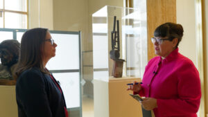 Two women stand in an art gallery, engaged in conversation. One is holding a notepad and pen. A sculpture is displayed in the background.