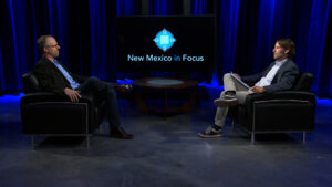 Two men are seated in armchairs facing each other in a studio with a screen displaying "New Mexico in Focus.