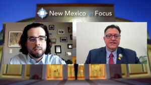 Two men in formal attire are on a split-screen video call with a "New Mexico in Focus" logo above. The background shows a blurred image of a building entrance.