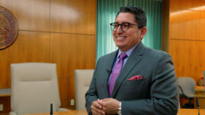 A man in a suit and tie stands smiling in a wood-paneled office with conference chairs and a seal on the wall.