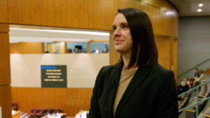 A woman in a blazer stands indoors, looking to the side. A wooden-panelled room with people and screens are visible in the background.