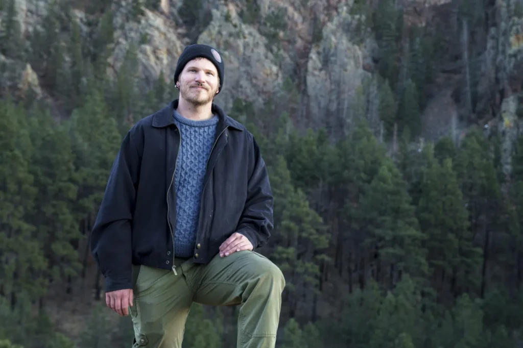 Person in a beanie and jacket poses with one knee up against a backdrop of pine trees and rocky cliffs.