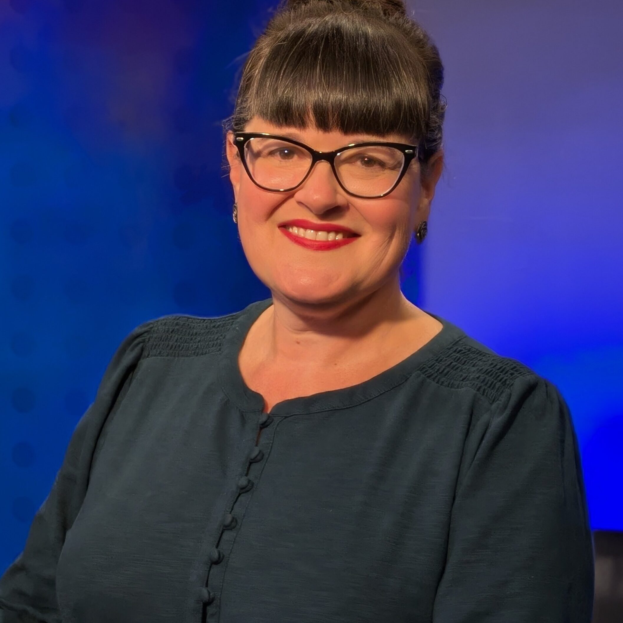 A person with glasses and a bun hairstyle smiles at the camera. They are wearing a dark green blouse, standing against a blue background.