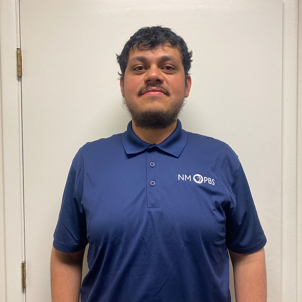 Man in a blue polo shirt with "NM PBS" logo stands against a white door background.