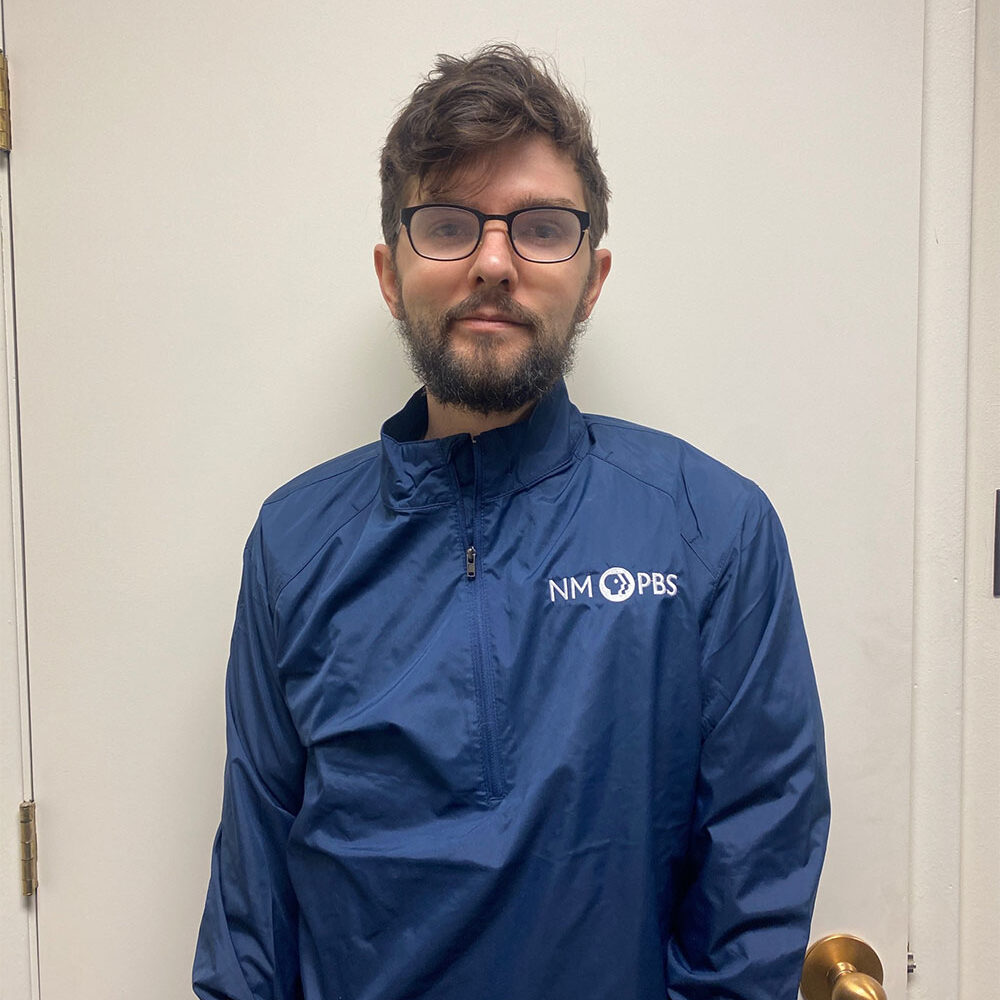 A person with glasses and a beard stands against a white door, wearing a blue NM PBS jacket.