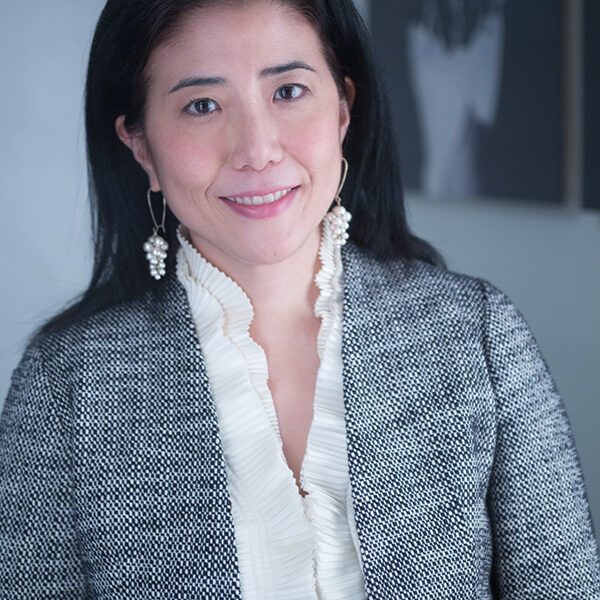 A Chinese-American woman with long dark hair is wearing a gray patterned blazer, a white ruffled blouse, and drop earrings. She is smiling while standing in front of the blurred backdrop of an Albuquerque film festival event.