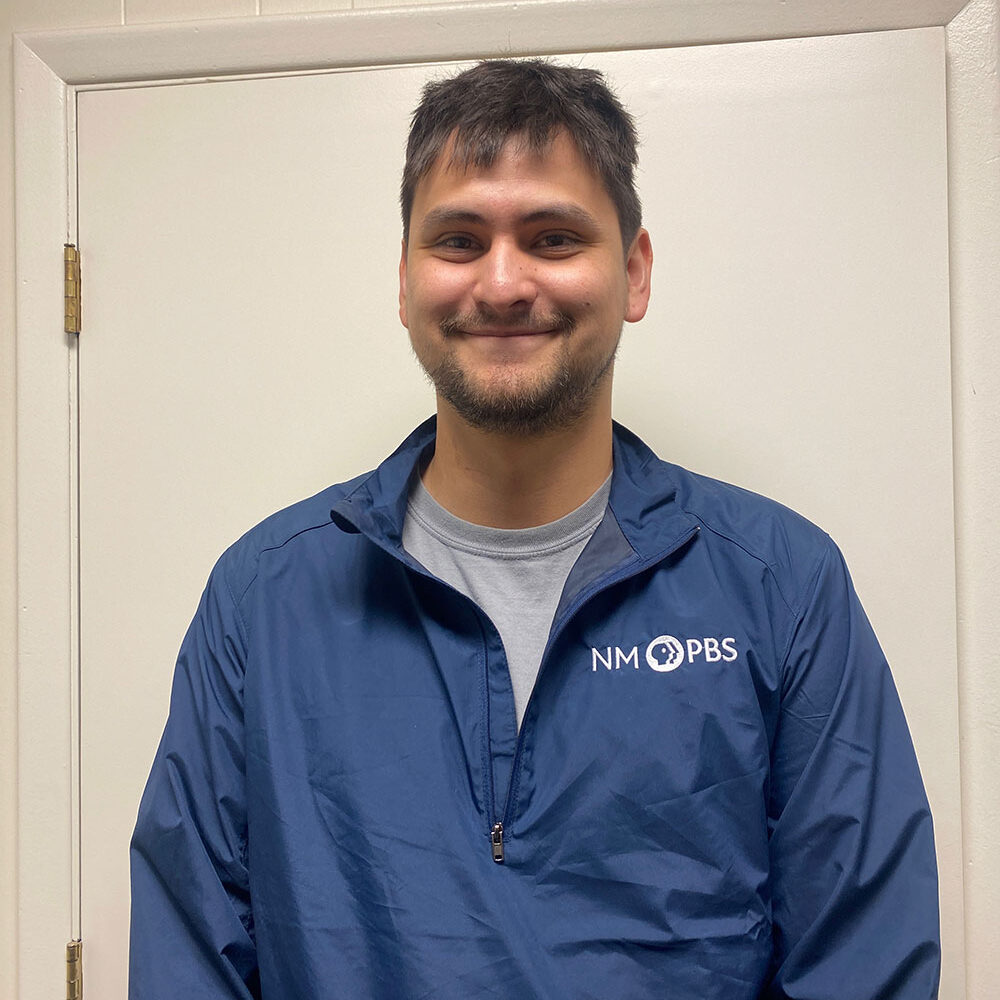 Person smiling, wearing a blue NM PBS jacket, standing in front of a white door.