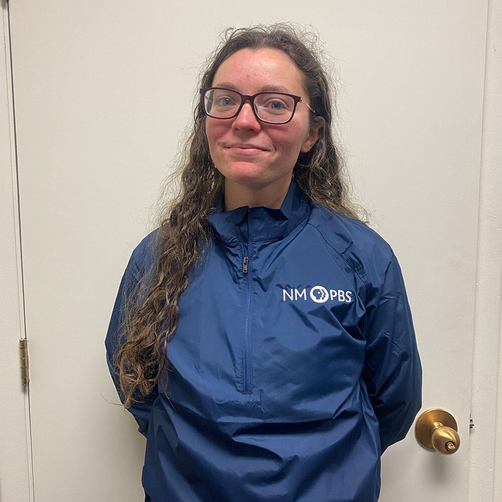 Person wearing glasses and a blue NM PBS jacket stands in front of a white door, smiling slightly.