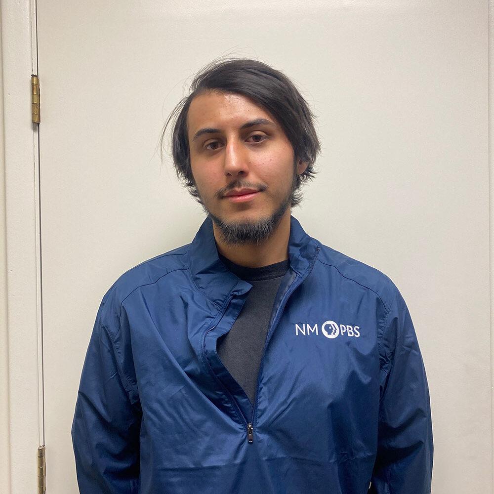 A person with dark hair and a beard stands in front of a white door, wearing a blue NM PBS jacket and a black shirt.