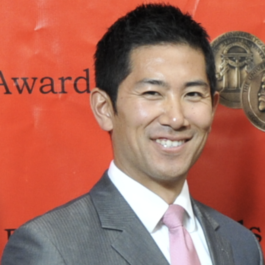 A Chinese-American man in a grey suit with a pink tie is smiling in front of a red backdrop with black text and a circular emblem at the Albuquerque Film Festival.