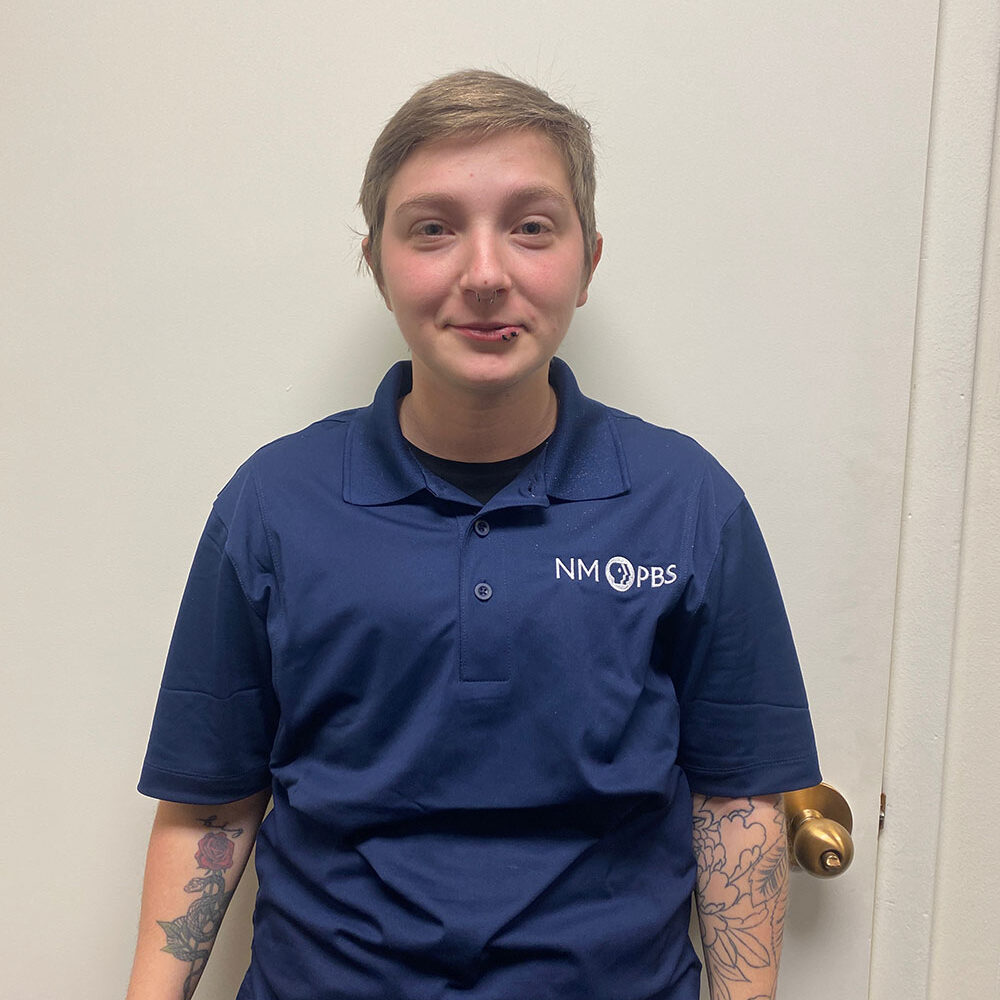 Person with short hair in a blue NM PBS polo shirt stands in front of a white door, with tattoos visible on both forearms.