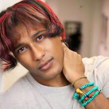 A person with medium-length red-tinted hair, reminiscent of a rising star at the Albuquerque Film Festival, touches their neck while wearing beaded bracelets and a light-colored shirt.