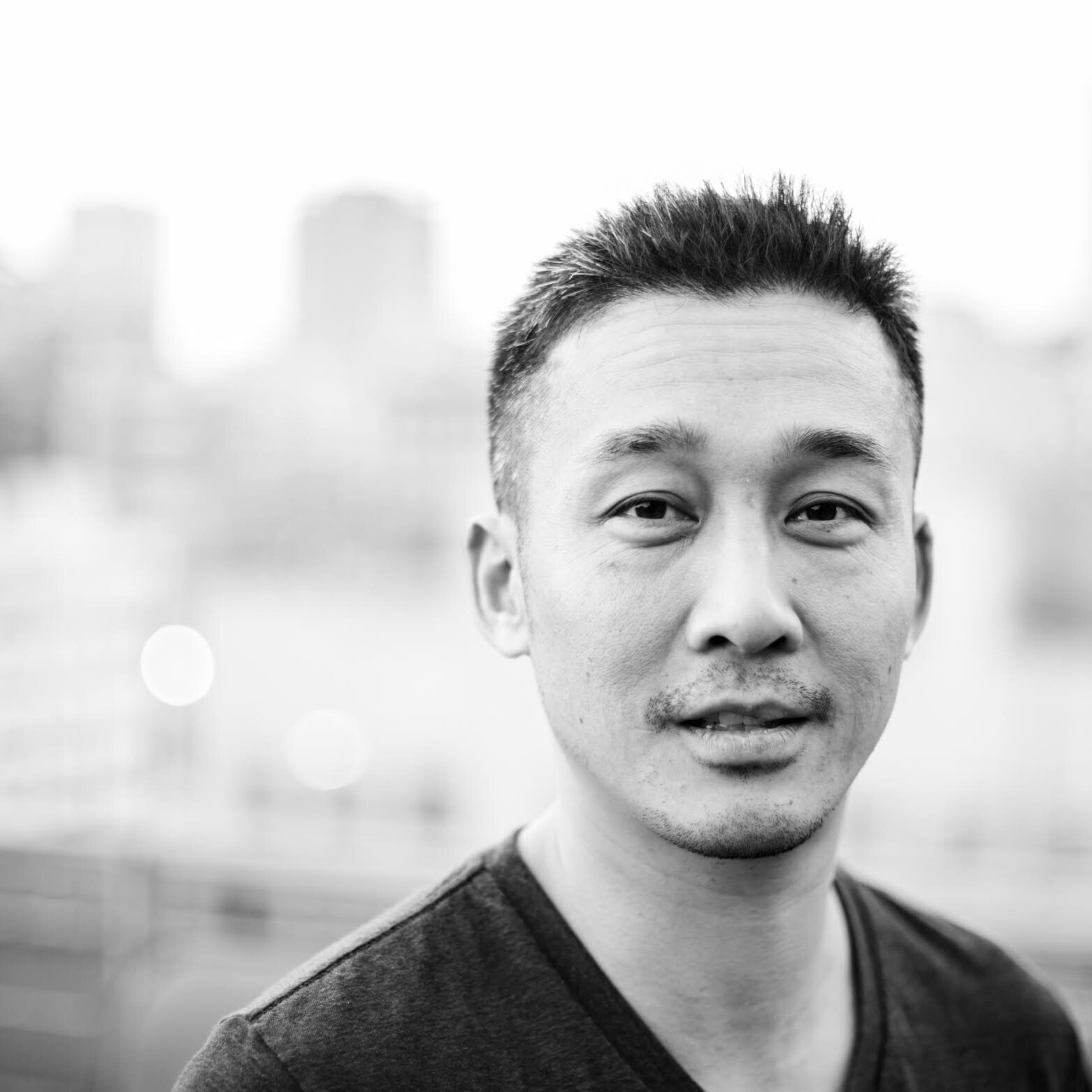 Black-and-white photo of a Chinese-American man with short hair and a slight smile, standing outdoors with a blurred urban background in Albuquerque.